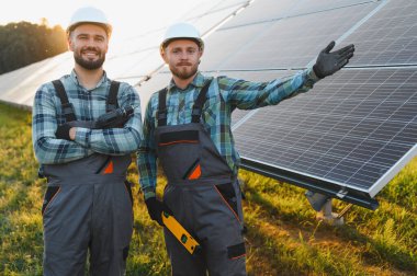 Bir güneş çiftliğinde duran iki mühendis, biri matkap tutuyor, diğeri yenilenebilir enerji altyapısını işaret ediyor.