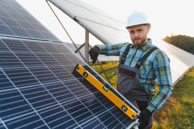 Sert şapka takan ve iş üniforması giyen adam güneş paneli kuruyor, bir seviye aracı ile hizalamayı kontrol ediyor, sürdürülebilir gücü destekliyor