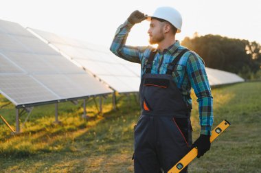 Yenilenebilir enerji işçisi sabit bir şapka ve tulum giyiyor, ruh seviyesini koruyor, güneş batarken bir güneş çiftliğinde duruyor.