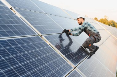 Sürdürülebilir bir enerji santraline güneş panelleri kuran adam, güneş ışığı altında fotovoltaik diziyi koruyan baret ve tulumlu teknisyen.