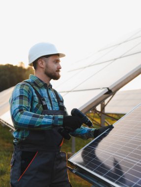Erkek işçi sert bir şapka ve eldiven takıyor, güneş panellerini matkapla birleştiriyor, temiz enerji üretiyor.