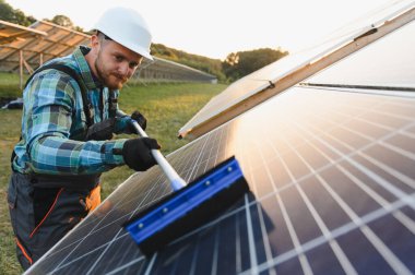 Sert şapkalı ve iş elbiseli bir adam yenilenebilir enerji için bakım çalışmaları sırasında güneş panelini temizliyor.