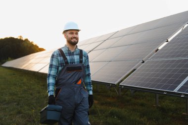 Şapkalı ve tulumlu gülümseyen teknisyen gün batımında bir dizi güneş panelinin yanında duran bir alet çantası tutuyor.