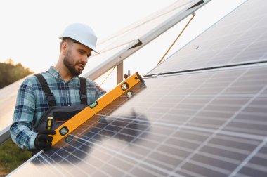 Sert şapkalı teknisyen, güneş panellerinin yenilenebilir enerji çiftliği ve açık hava pv kurulumundaki hizalanmasını kontrol etmek için ruh seviyesini kullanıyor.