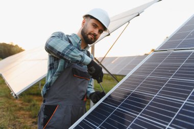 Sert şapkalı erkek teknisyen güneş panelini tornavidayla kuruyor. Gün batımında yenilenebilir enerji çiftliğinde çalışıyor.