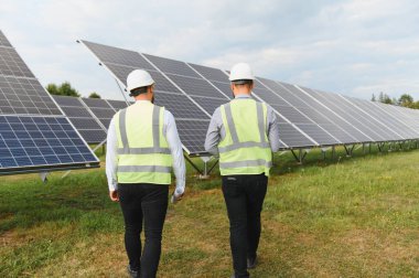 Güvenlik donanımlı iki mühendis, yenilenebilir enerji, sürdürülebilirlik ve temiz teknolojiyi sembolize ederek büyük güneş paneli kurulumunu denetliyor.