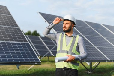 Güneşli bir fotovoltaik çiftlikteki güneş panellerini incelerken, sürdürülebilir enerji gelişimi planlarken, sabit şapka ve güvenlik yeleği mühendisinin elinde planlar bulunuyor.
