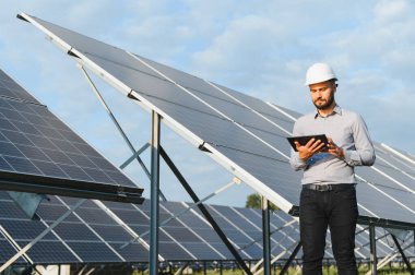 Sert başlıklı mühendis, berrak mavi gökyüzünün altında yenilenebilir enerji odaklı tablet kullanarak büyük bir güneş çiftliğindeki sıra sıra güneş panellerini inceliyor.