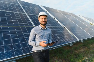 Güneş panelleri önünde duran sert şapkalı ve tabletli erkek mühendis, güç üretimini ve sürdürülebilirliği izliyor.