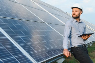 Güneş çiftliğindeki sıra sıra güneş panellerini incelerken elinde tablet bulunan erkek mühendis temiz enerji ilerlemesini simgeliyor.