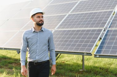 Güneş çiftliğinde yürüyen, kasklı bir erkek uzman güneşli bir günde yenilenebilir enerji tesisatını kontrol ediyor.