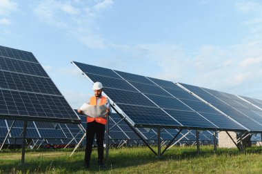 Güvenlik yeleği giyen mühendis ve güneş enerjisi santralinin planlarını gözden geçirip yenilenebilir enerji üretimini garanti altına alıyor.