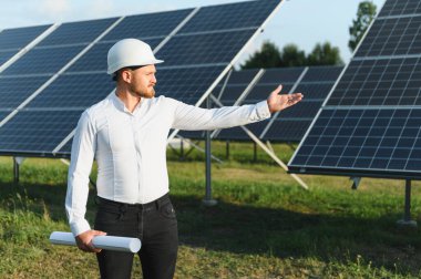 Mühendis sabit bir şapka takıyor ve elinde bir plan tutuyor. Yenilenebilir enerji çiftliğinde bir dizi güneş paneli sunuyor.