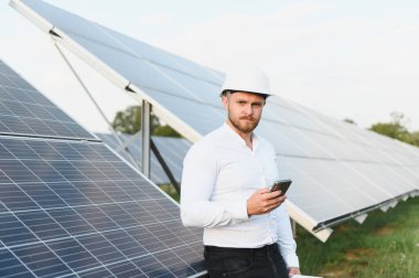 Kasklı mühendis güneş paneli alanını inceliyor, elinde akıllı telefon var. Yenilenebilir enerji teknolojisi ve sürdürülebilir güç sunuluyor