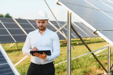 Bir elektrik santralinde güneş paneli sıralarını incelerken sabit şapka takan ve dijital tablet tutan mühendis