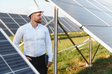 Erkek mühendis, başlık takıyor ve elinde dijital tablet tutuyor. Gündüz vakti bir çiftlikte güneş paneli kurulumunu yönetiyor.
