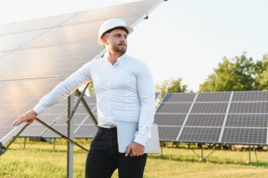 Sert bir şapka takan ve dizüstü bilgisayar tutan mühendis güneş paneli kurulumunu kontrol edip yenilenebilir enerjiye odaklanıyor.