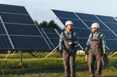Başlık takan ve üniforma giyen iki mühendis yeşil bir alandaki güneş enerjisi santralini kontrol ediyor ve tartışıyorlar.