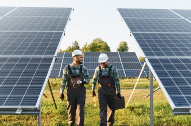 Güneş paneli alanında yürüyen ve konuşan, yeni sürdürülebilir enerji kaynağı kuran, sert şapkalı ve tulumlu mühendisler.