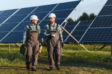 Sert şapka ve tulumlu mühendisler güneş paneli alanından geçerek yenilenebilir enerji kurulum planlarını tartışıyorlar.