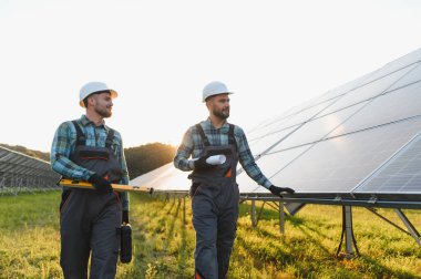 Güneş enerjisi işçileri güneş enerjisi tarlalarında yürüyorlar, ellerinde planlar ve araçlar var, yenilenebilir enerji üretimini sağlıyorlar.