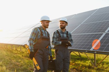 İki mühendis, büyük bir güneş paneli kurulumunun önünde dikilirken, başlık ve tulum giyip tartışıyorlar.
