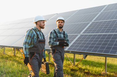 Güneş çiftliğinde yürüyen güvenlik ekipmanlı iki mühendis sürdürülebilirlik için çevre dostu alternatif enerjiyi kontrol ediyorlar.