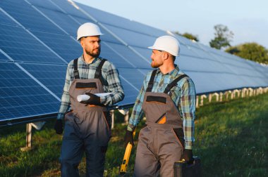 Güneş enerjisi santralinde yürüyen ve tartışan iki mühendis ellerinde planlar ve aletler tutuyorlar.