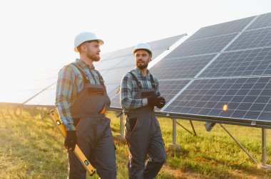 Sert şapka ve tulum giyen iki teknisyen bir dizi güneş paneli kuruyor, yenilenebilir enerji kullanıyorlar.