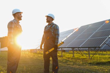 Gün batımında güneş panellerinin yanındaki bir tarlada duran kasketli iki gülümseyen mühendis temiz enerji üzerinde çalışıyorlar.