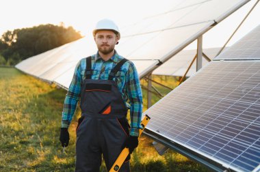 Siyah şapkalı ve üniformalı erkek işçi elinde aletlerle yeşil enerji alanına güneş panelleri yerleştiriyor.