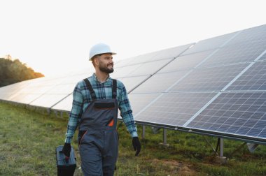 Genç teknisyen alet çantasıyla yürüyor, yenilenebilir enerji istasyonundaki güneş panellerini izliyor.