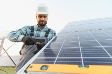 Güvenlik kaskı ve tulum giyen erkek işçi çelik çerçeveye fotovoltaik panel yerleştiriyor.