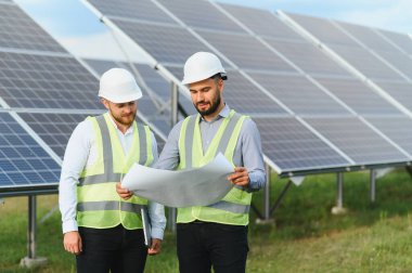 Güvenlik dişlisi olan iki erkek mühendis güneş panelleri önündeki planları gözden geçirir, takım çalışmasını, yeniliği ve temiz enerjiyi sembolize ederler.