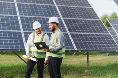 Geniş güneş panelleri önünde dizüstü bilgisayarda yenilenebilir enerji ilerlemesini sembolize eden koruyucu dişli inceleme planları yapan iki mühendis.