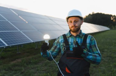 Baş parmaklarını gösteren ve güneş panellerinden gelen temiz enerjiyi sembolize eden parlayan bir ampul tutan mühendis.