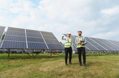 Güneş panellerinin yanında duran güvenlik yelekleri ve kasklı iki erkek mühendis dışarıdaki yenilenebilir enerji üretim alanını inceliyor.