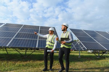 Güvenlik dişlisi iki mühendis güneş panellerini inceleyerek yenilenebilir enerji stratejileri ve sürdürülebilir enerji gelişimini tartışıyor.