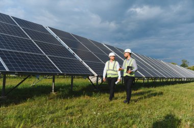 Güvenlik teçhizatı takan iki mühendis, yenilenebilir teknoloji ve sürdürülebilirliği sembolize ederek temiz bir enerji çiftliğinde güneş panellerini denetliyor.