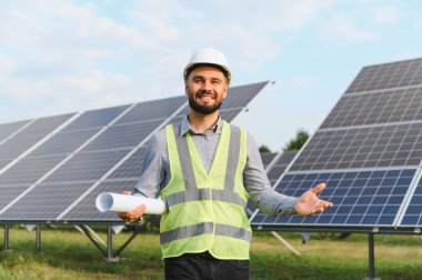 Kasketli ve güvenlik yeleği giymiş gülümseyen adam elinde planlar, büyük güneş paneli alanının önünde duruyor.