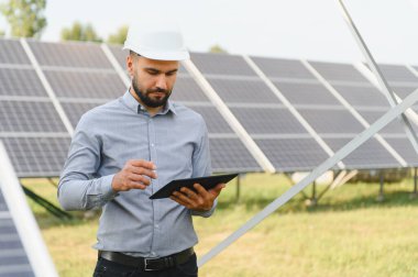 Dijital tablet kullanarak sabit şapka takan mühendis yenilenebilir enerji çiftliğindeki güneş panellerini izliyor.