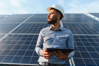 Sert şapkalı erkek mühendis bir tablet tutuyor, güneş panellerini inceliyor ve sürdürülebilir enerji için açık hava enerji sistemlerini izliyor.