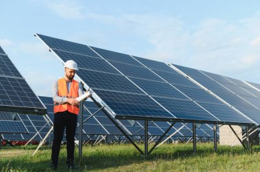 Kask ve güvenlik yeleği giyen mühendis, büyük bir güneş enerjisi santralinin planlarını kontrol ediyor, sürdürülebilirlik çabalarına katkıda bulunuyor.