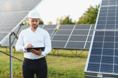 Kask ve beyaz gömlek giyen adam güneş paneli performansını güneş çiftliğinde dijital tabletle kontrol ediyor.