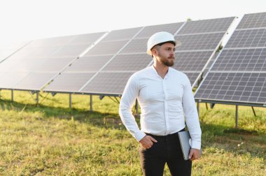 Sert şapkalı mühendis elinde dizüstü bilgisayarla açık bir gökyüzünün altında büyük bir güneş paneli alanı gözlüyor.