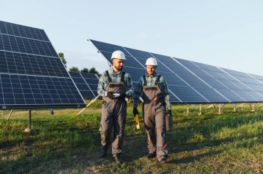 İki işçi güneş çiftliğinde yürüyor, bakım ve yenilenebilir enerji çözümlerini tartışıyorlar.
