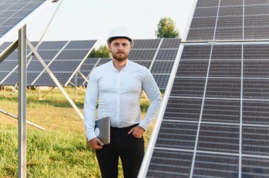 Genç mühendis sabit şapka takıyor ve dizüstü bilgisayar tutuyor, güneş paneli açıklarında duruyor, yeşil enerjiye odaklanıyor.