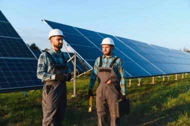 Güneş panellerinin önünde duran iki mühendis, ellerinde araçlar, yenilenebilir enerji üretimini denetliyorlar.