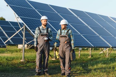 Sert şapkalı ve tulumlu iki işçi güneş enerjisi panellerini kontrol ederek temiz elektrik ve yenilenebilir enerji sağlıyor.