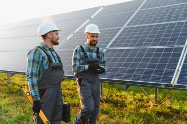İki mühendis, yeşil bir alanda güneş paneli kurulumu üzerinde çalışırken, sıkı şapkalar ve tulumlar giyip tartışıyorlar.